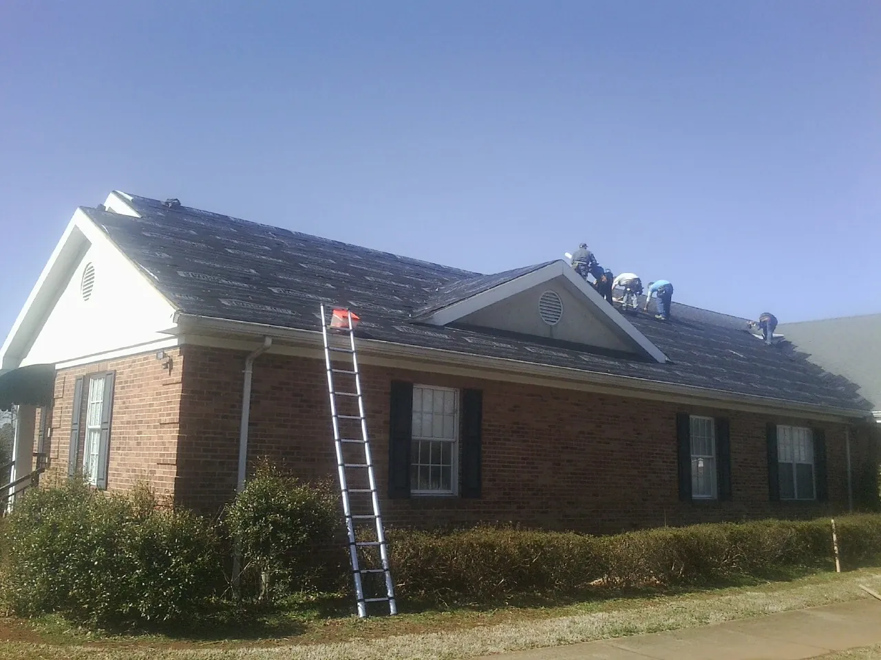 Crew tearing shingles off a brick ranch roof with a ladder set — Emergency Roof Repair work by our crew in Westhampton Beach
