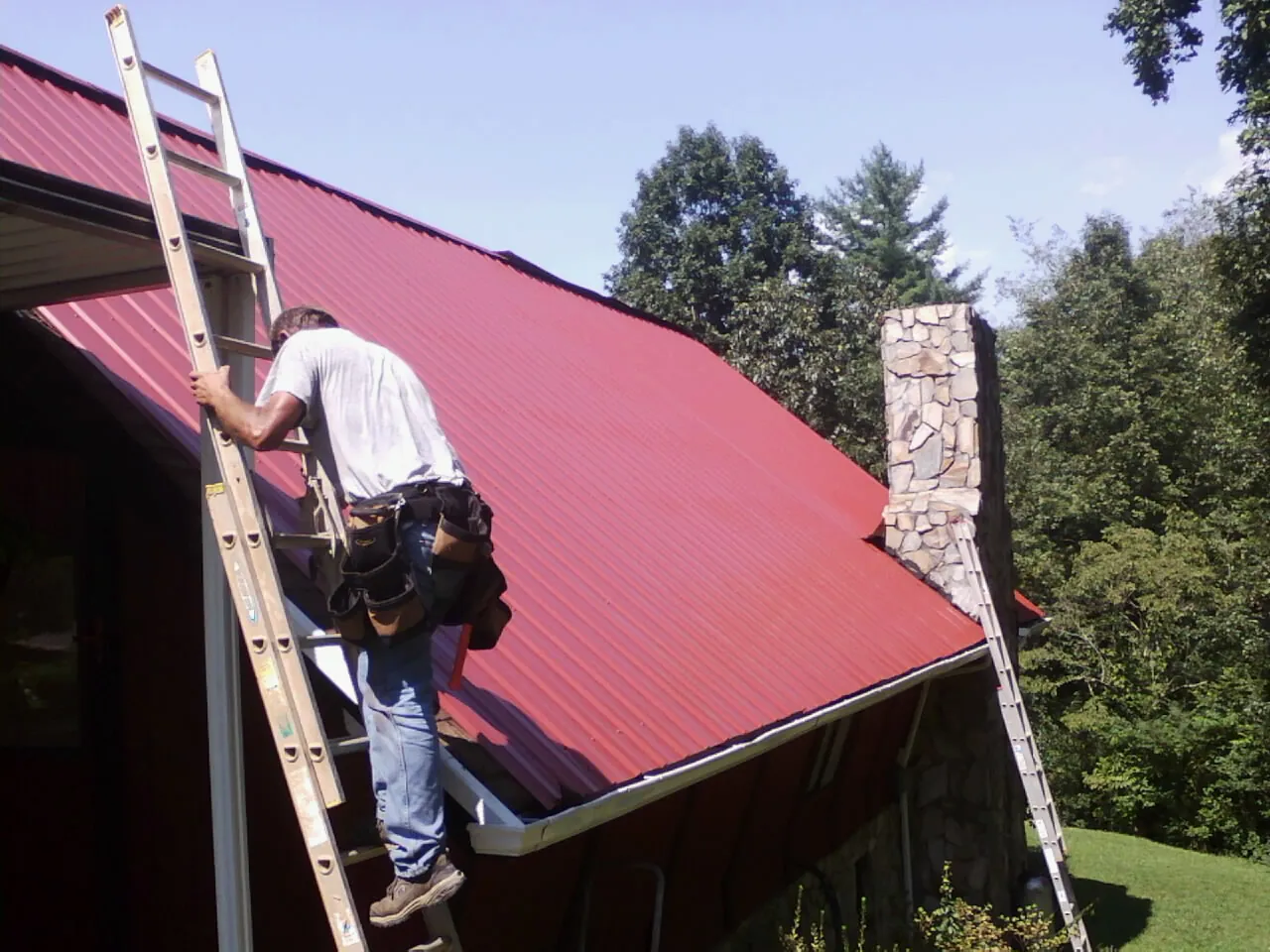 Roofer on a ladder beside a red metal roof and stone chimney — Soffit & Fascia Repair by Roofing Brothers in Westhampton Beach